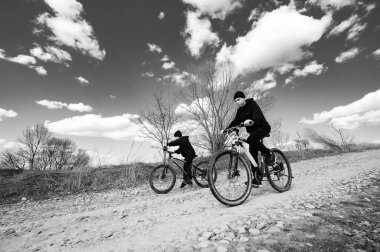 Çakıl yolunda bisiklete binen iki kişinin siyah beyaz fotoğrafı. Gökyüzü parçalı bulutlu. Çıplak ağaçlar yol boyunca diziliyor. Huzurlu ve huzurlu bir sahne.