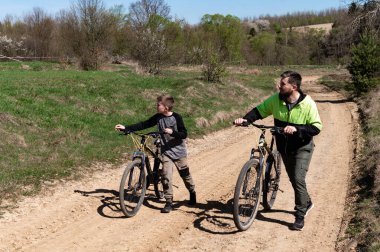 Güneşli bir günün tadını çıkaran bir adam ve çocuk yemyeşil çayırların ve ağaçların arasında kirli bir patika boyunca bisikletlerini yürüyorlar. Aile eğlencesi için iç açıcı bir sahne..