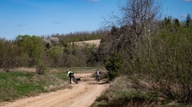 İki bisikletçi ve bir köpek ağaçlık bir alanda kumlu bir yolda görülmüş. Gün güneşli ve doğal ortam huzur dolu. Sahne, eğlence amaçlı bisiklet sürmeyi öneriyor..