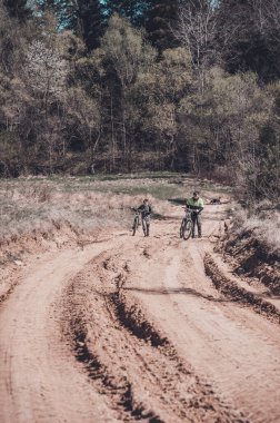 İki figür, muhtemelen baba ve oğul, kumlu bir patika boyunca bisikletlerini yürütürler. Ağaçlar yol boyunca uzanır, güneş ışığı yaprakların arasından süzülür. Barışçıl bir açık hava sahnesi.