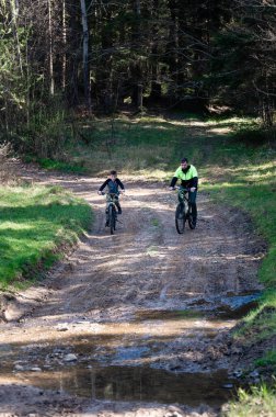Bir adam ve bir çocuk ağaçlık bir alanda çamurlu bir patika boyunca bisiklet sürüyorlar. Güneş ışığı ağaçların arasından süzülüp parlak ve maceralı bir sahne yaratır..
