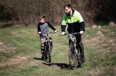Bir adam ve çocuk çamurlu bir yolda bisiklet sürüyor, güneşli bir günün tadını çıkarıyorlar. Açık araziler ve mavi gökyüzü huzurlu bir manzara yaratır. Dağ bisikleti sürüyorlar..