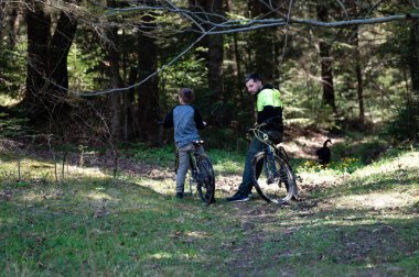 Bir adam ve bir çocuk, huzurlu ormanlık araziyi almak için bisikletlerini durdurur. Güneş ışığı ağaçların arasından süzülüp sakin bir atmosfer yaratır..