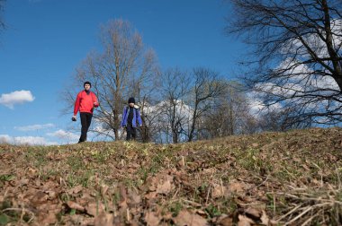 İki figür, bir yetişkin ve bir çocuk, açık mavi gökyüzünün altında yapraklarla kaplı zemin ve çıplak ağaçlarla çevrili bir tepenin üzerinde bir yol boyunca yürürler..
