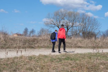 Sırt çantalı iki genç, kır yolunda yürüyorlar. Gökyüzü parlak mavi ve çevresindeki bitki örtüsü kuru ve kahverengidir. Yakınlarda bir ağaç duruyor..