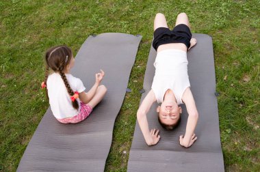 İki çocuk, çimenli bir bölgedeki gri paspaslar üzerinde yoga seansının keyfini çıkarıyor. Bir kız meditasyon pozunda otururken bir erkek arkaya doğru eğiliyor. Zindelik ve refahı teşvik eden bir sahne.