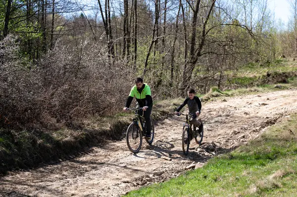 İki bisikletçi, bir yetişkin ve bir çocuk, kırsal bir yolda dağ bisikletlerine biniyorlar. Güneş ışığı ağaçların arasından akar, gölgeler bırakır. Barışçıl bir açık hava macerası.