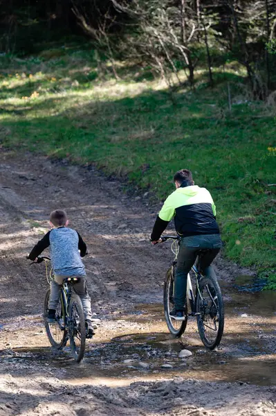 İki bisikletçi, bir yetişkin ve bir çocuk ormanda çamurlu bir patika boyunca dağ bisikleti sürüyor. Güneş ışığı ağaçların arasından süzülür, patikada benekli gölgeler yaratır..