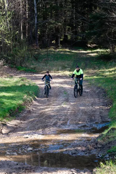 Bir adam ve bir çocuk ağaçlık bir alanda çamurlu bir patika boyunca bisiklet sürüyorlar. Güneş ışığı ağaçların arasından süzülüp parlak ve maceralı bir sahne yaratır..