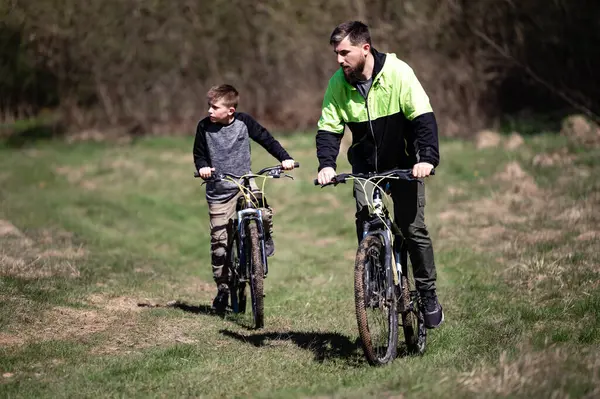 Bir adam ve çocuk çamurlu bir yolda bisiklet sürüyor, güneşli bir günün tadını çıkarıyorlar. Açık araziler ve mavi gökyüzü huzurlu bir manzara yaratır. Dağ bisikleti sürüyorlar..