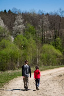 İki çocuk, bir oğlan ve bir kız, kırsal bir yolda geziniyorlar. Arka planda canlı bir bahar ormanı görülüyor. Huzurlu ve doğal bir ortam yaratıyor..