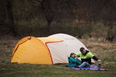Bir çift, ağaçlarla çevrili çimenli bir düzlükte, açık sarı çadırlarında, sıcak içkilerini yudumlayarak ve dışarıdaki sessizliğin tadını çıkararak huzurlu bir anın tadını çıkarıyor..