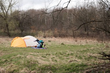 İki kampçı çadırlarını, altında yumuşak çimenler ve yukarıda bulutlu gökyüzü olan sakin bir orman açıklığında hazırlıyorlar. Doğa onları çevrelemiş, açık havanın güzelliğini sergiliyor..
