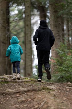 Doğada paylaşılan sıcak anlar parlak ceketli bir çocuk olarak ve sakin bir orman yolunda, yüksek ağaçlar ve yumuşak toprakla çevrili bir yetişkin yürüyüşü olarak gözler önüne seriliyor..