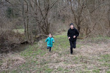 Çocuklar, çıplak ağaçlarla çevrili, yumuşak bir derenin yanında çimenli bir patika boyunca koşar. Kahkahaları, sakin kış manzarasını keşfederken havayı dolduruyor..