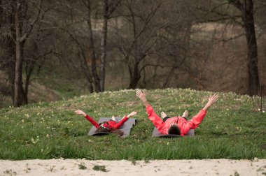 Ağaçlarla çevrili yemyeşil bir alanda yoga yapan iki insan öğleden sonra güneşinin rahatlatıcı kucağında güneşlenirken bedenleri ve zihinleri uyumlu hale getiriyorlar..