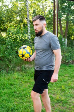 Spor kıyafetli sakallı bir adam bir futbol topu tutuyor. Çimenli bir alanda duruyor, muhtemelen bir park ya da eğlence alanında. Ortam açık ve doğal..