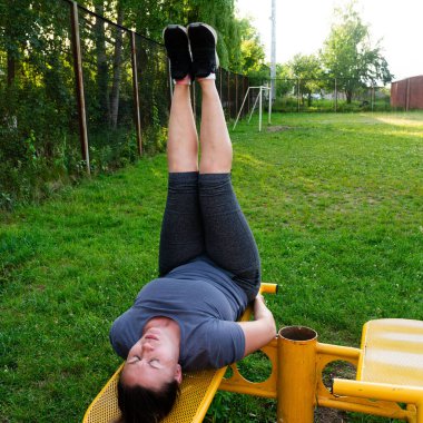 Açık hava egzersiz bankında bacak kaldıran bir kadın. Sahne çimenli bir park alanı. Atletik kıyafetler giyiyor..