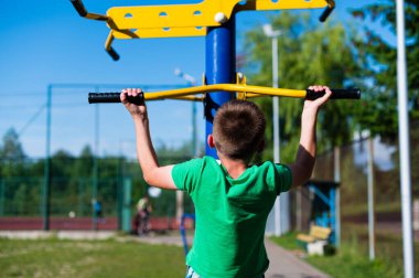 Genç bir çocuk açık hava egzersiz makinesinde barfiks çekiyor. Güneşli gün ve yeşil çevre pozitif ve aktif bir atmosfer yaratır..