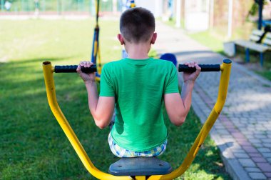 Genç bir çocuk vücut gücüne odaklanmak için açık hava spor aletleri kullanıyor. Güneşli ortam halka açık bir park ya da eğlence alanı öneriyor.