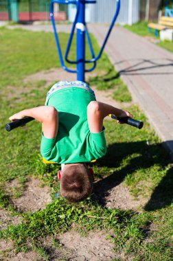 Genç bir çocuk açık hava spor aletlerine baş aşağı sarkıyor. Sahne güneşli ve açık hava, eğlenceli ve aktif bir gün olduğunu gösteriyor..