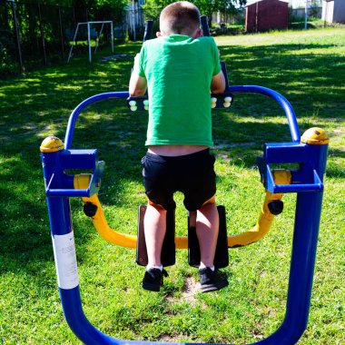 Genç bir çocuk açık havada bacak pres makinesi kullanıyor. Güneşli park ortamı sağlık ve sağlığa odaklandığını gösteriyor. O yeşil bir gömlek ve koyu şort giyiyor..