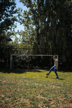 Bir genç güneşli bir öğleden sonranın tadını çıkarır, futbol yeteneklerini açık havada çalışır. Yeşillik ve ağaçlarla çevrili bir kaleye doğru topa vuruyor..