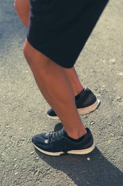 Bir insanın bacaklarının ve ayaklarının koşu ayakkabılarının içinde asfalt bir yolda esnediği yakın plan. Güneş gölgeler bırakır, sahnenin atletik havasını güçlendirir..