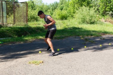 Genç bir atlet çeviklik tatbikatı yapıyor. Çimenli bir bölgenin yakınındaki asfalt bir yüzeye kurulmuş sarı koniler etrafında yoğun bir şekilde hareket ediyor. Ayar güneşli bir günde açık havada..