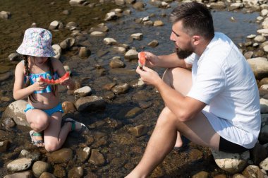 Bir adam ve genç bir kız, her ikisi de yaz kıyafetleri içinde, pürüzsüz nehir kayalarının arasında oturup, mutlu bir şekilde karpuz dilimleri yiyorlar. Sığ, berrak su etraflarında yavaşça akıyor..