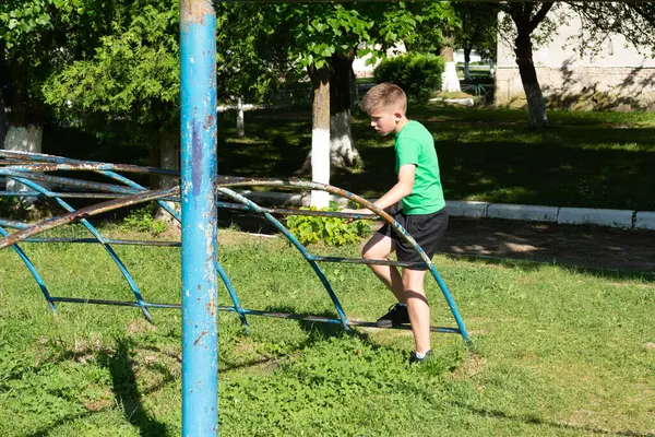 Genç bir çocuk dışarı çıkan yıpranmış mavi bir metale tırmanıyor. Sahne yemyeşil ağaçları olan çimenli bir park. Güneş ışığı sahneyi aydınlatır..