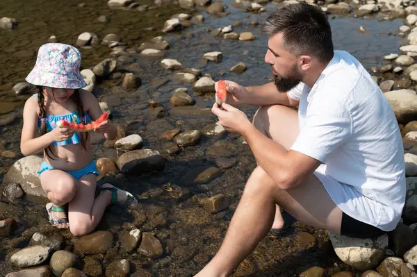 Bir adam ve genç bir kız, her ikisi de yaz kıyafetleri içinde, pürüzsüz nehir kayalarının arasında oturup, mutlu bir şekilde karpuz dilimleri yiyorlar. Sığ, berrak su etraflarında yavaşça akıyor..