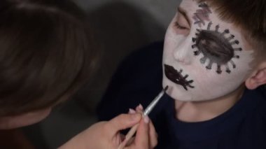 Bir kadının genç bir çocuğa yüz boyası uygulayarak yüzünü detaylı bir kafatası tasarımına dönüştürdüğü yakın plan. Sahne samimi ve paylaşılan bir yaratıcılık anı gösteriyor..