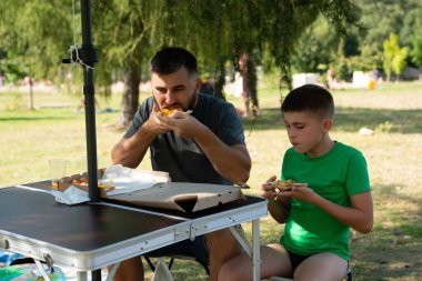 Bir adam ve bir çocuk parkta küçük bir masada oturup pizza yiyorlar. Güneşli bir günde, bir ağacın gölgesinde gündelik bir yemeğin tadını çıkarıyorlar..