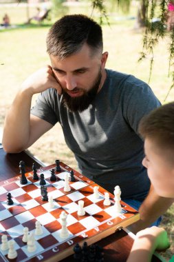 Satranç tahtasına odaklanmış sakallı bir adam ağaç gölgesinde kendinden genç biriyle satranç oynuyor. Sahne huzurlu ve güneşli, sakin bir öğleden sonranın göstergesi..
