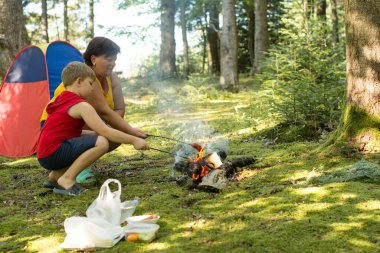 Bir kadın ve genç bir çocuk yemyeşil bir ormanda kamp ateşi üzerinde yemek pişiriyorlar. Küçük bir çadırın yanında oturuyorlar, dışarıda huzurlu bir deneyimin tadını çıkarıyorlar..