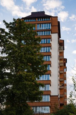 Çarpıcı bir modern bina, hareketli ağaçların arasından yükseliyor, büyük pencerelerinden yansıyan güneş ışığını yansıtıyor, hareketli bir şehirde doğanın ve çağdaş mimarinin bir karışımını sergiliyor..