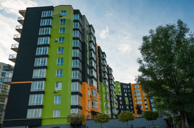 Parlak yeşil, turuncu ve gri renklerle boyanmış modern bir apartman kompleksi berrak bir gökyüzü altında yükselir, canlı bir mahallede yeşil ve kentsel canlılık ile çevrilidir..