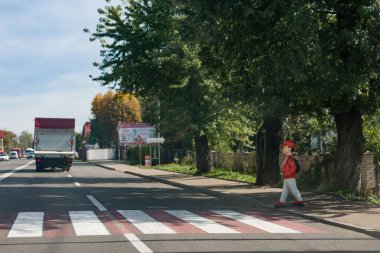 Sıradan giyimli bir birey yaya geçidinde yürüyor, ağaçlarla çevrili, bir kamyon geçerken, büyüleyici kasabada sakin bir atmosfer yaratıyor..