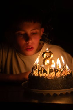 Genç bir çocuk heyecanla eğiliyor, nefis doğum günü pastasının mumlarını üflemek üzere, neşeli süslemelerle süslenmiş, bir neşe anı yaratıyor.