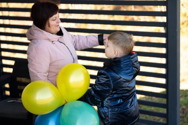 Bir büyükanne, elinde renkli balonlar tutan torunuyla sevgiyle etkileşime girer. Güneşli bir öğleden sonra gölgeli bir yapının altında keyifli bir anı paylaşıyorlar..