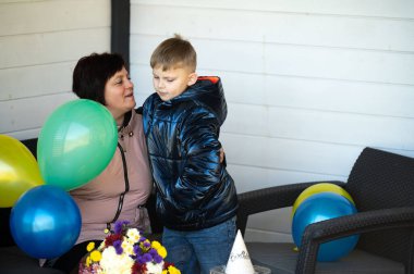 Sıcak bir an paylaşılırken bir anne, oğlunu canlı balonlar ve güzel bir çiçek aranjmanı arasında kucaklıyor ve açık havada neşeli bir atmosfer yaratıyor..