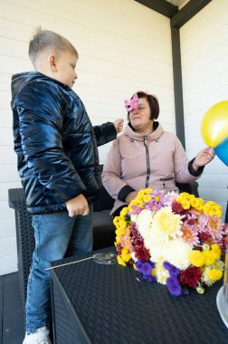 Bir çocuk neşeli bir hediye sunar canlı çiçeklerle çevrili bir kadına, açık mavi gökyüzü altında bir aşk ve mutluluk anı yakalar.