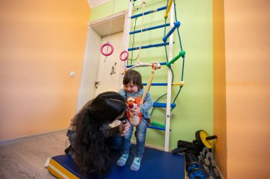 Bir çocuk renkli bir oyun odasında bir bakıcı tarafından tutulurken parlak bir şekilde gülümser. Tırmanış ekipmanları ve oyuncaklar etraflarını sararak oyun ve bağ kurmak için neşeli bir atmosfer yaratıyor..