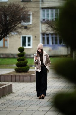 Sıcak bir şekilde giyinmiş bir kadın huzurlu bir yolda yürüyor, düzenli çalıların arasında telefonuna bakıyor ve sakin bir parkta sakin bir öğleden sonra..
