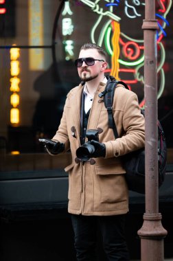 Şık bej bir palto giyen güneş gözlüklü genç bir adam kaldırımda güven içinde duruyor. Canlı şehir enerjisini fotoğraf makinesi ve telefonuyla neon tabelaları arasında yakalıyor..