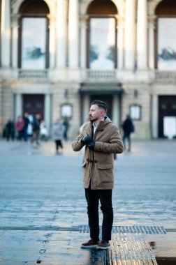 Şık bir palto giyen bir adam, canlı bir meydanda düşünceli bir şekilde duruyor, canlı kalabalığı inceliyor ve şehirde hareketli bir öğleden sonra boyunca büyüleyici bir mimariyi inceliyor..