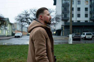 Bulutlu bir öğleden sonra, sıcak kahverengi bir ceket giymiş bir adam, çağdaş binalar ve iskelet ağaçlarla çerçevelenmiş yağmurla öpülmüş bir şehir parkında düşünceli bir şekilde yürür..