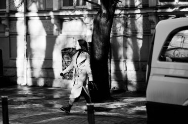 Zarif bir palto giymiş bir kadın büyüleyici bir sokakta yürüyor, ağaçların arasından süzülen benekli güneş ışığıyla aydınlatılıyor, mükemmel bir şehir ortamı yaratıyor..