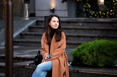 Parıldayan ışıklar ve yeşilliklerin ortasında genç bir kadın, şık bir paltoyla serin akşam havasına sıcak bir şekilde sarılan taş bir bankta sessiz bir anın tadını çıkarıyor..